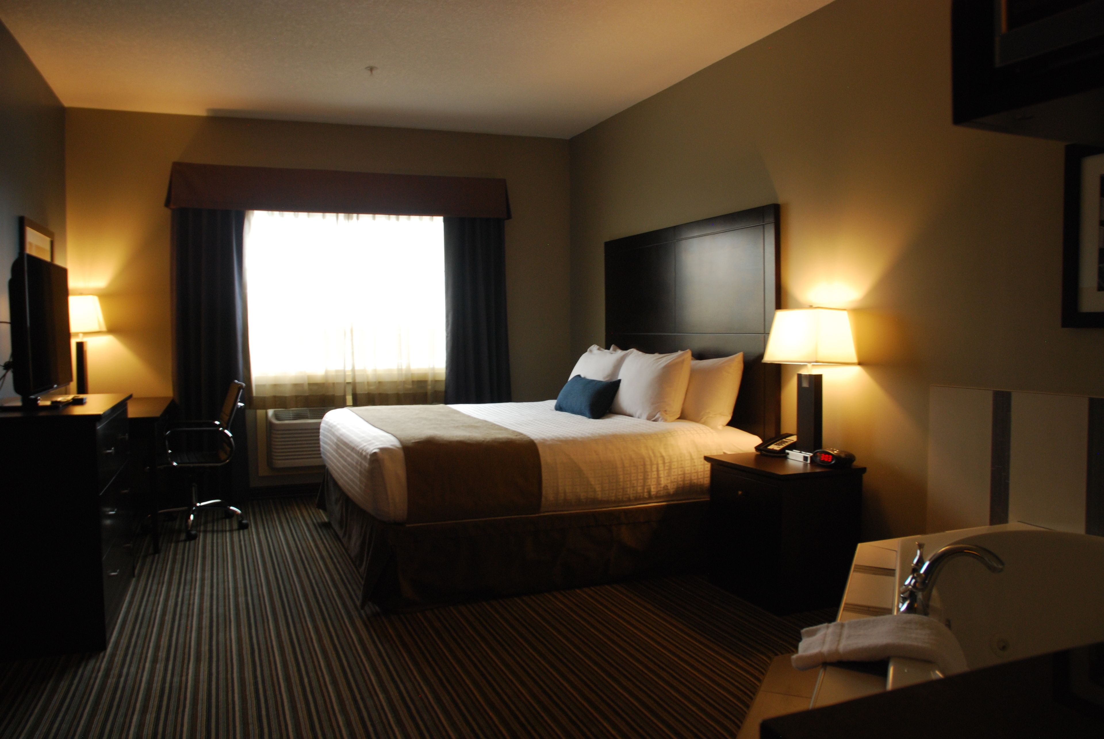 Une chambre d’hôtel comprenant un lit, des lampes de chevet, un bureau avec une chaise, une télévision et une salle de bain avec un lavabo et un miroir.