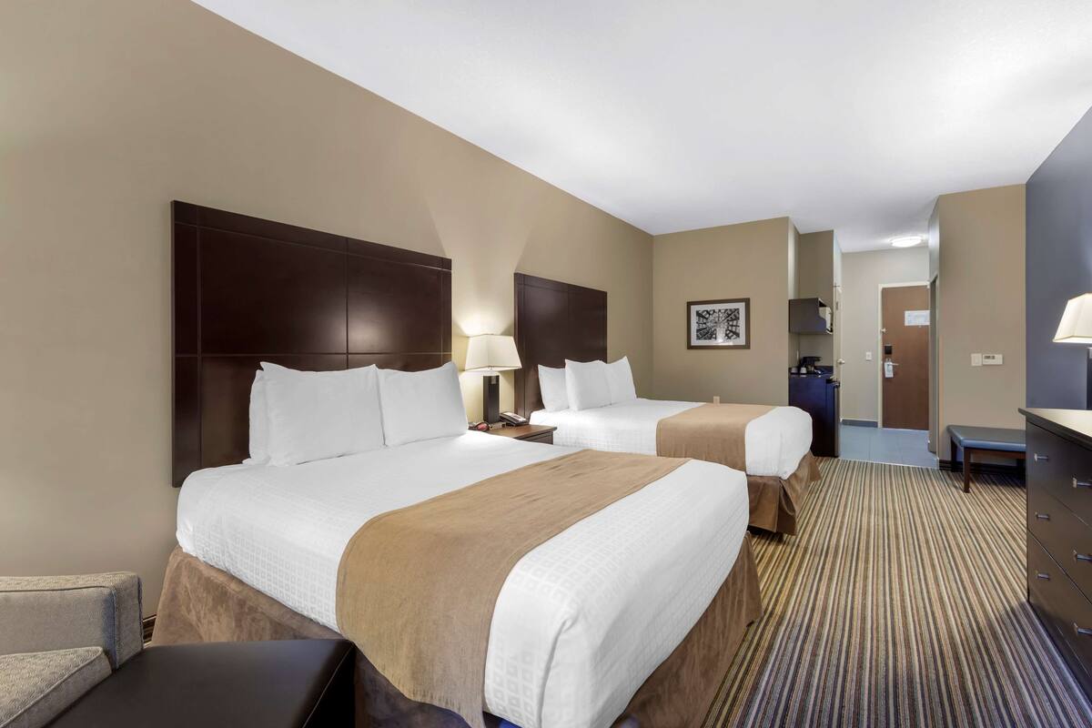 A hotel room with two beds, a large dark headboard, bedside lamps, a desk, and a bench.