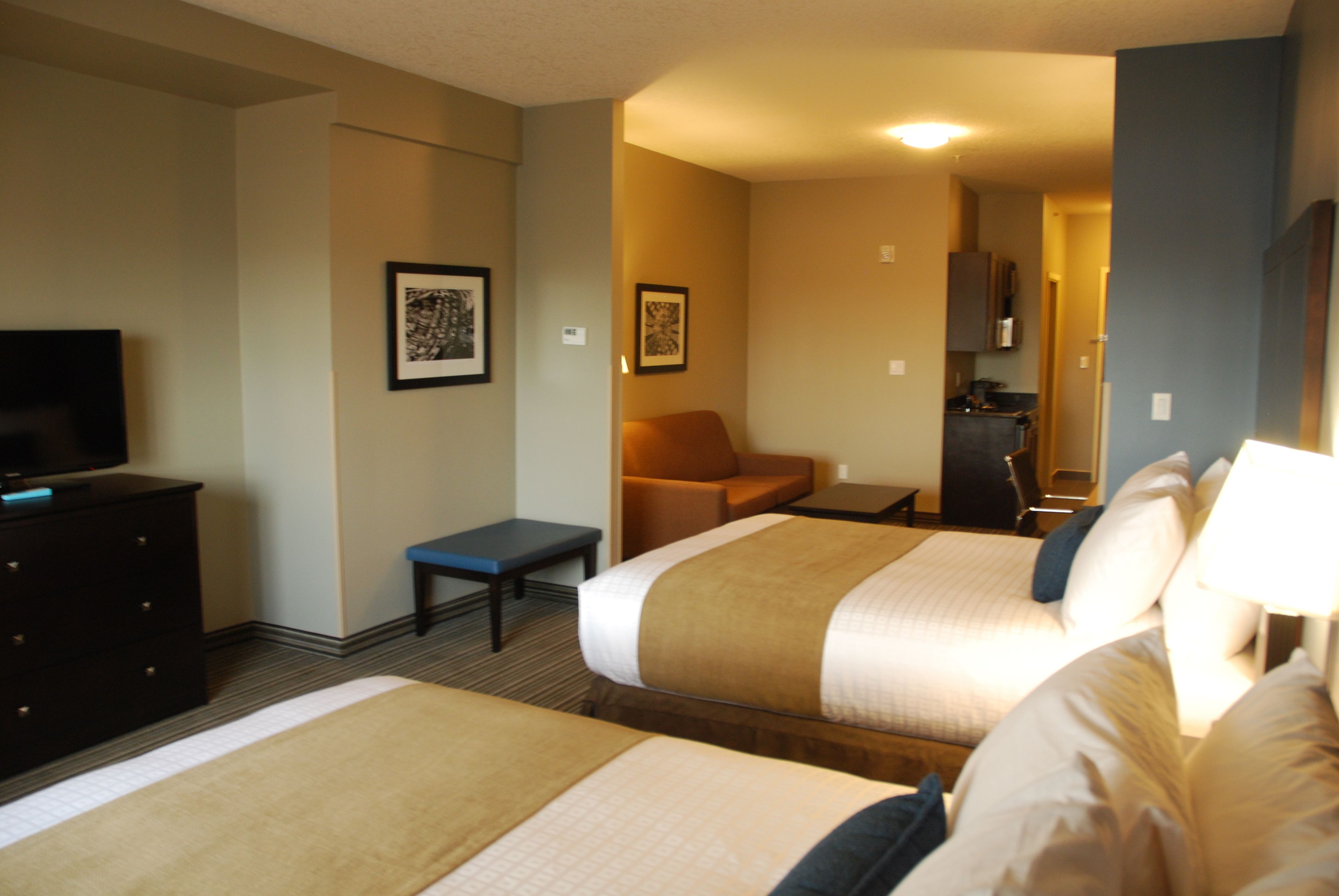 A hotel room with two beds, a desk, a chair, a television, and a kitchenette.