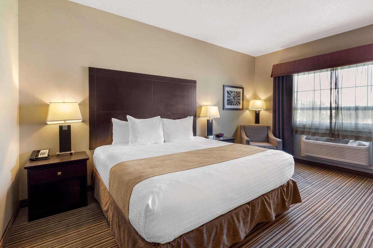 A hotel room with a large bed, two bedside tables with lamps, a chair, and a large window with curtains.