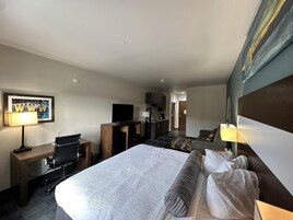 A hotel room with a large bed, a desk with a computer, a television, and a kitchenette.