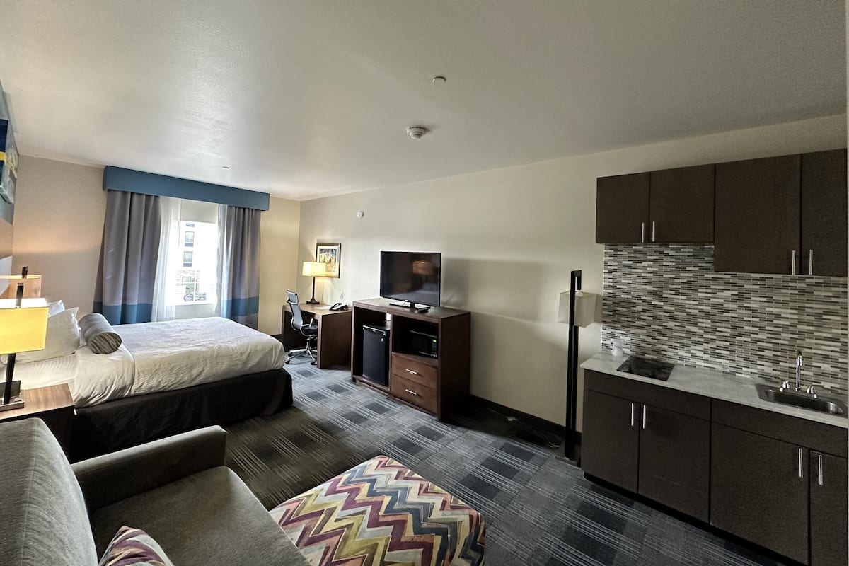 A hotel room with a bed, a desk with a computer, a TV, a kitchenette, and a sofa.