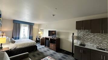 A hotel room with a bed, a desk with a computer, a TV, a kitchenette, and a sofa.
