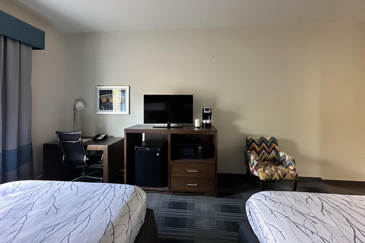 A hotel room with two beds, a desk with a TV, a coffee maker, and a patterned armchair.