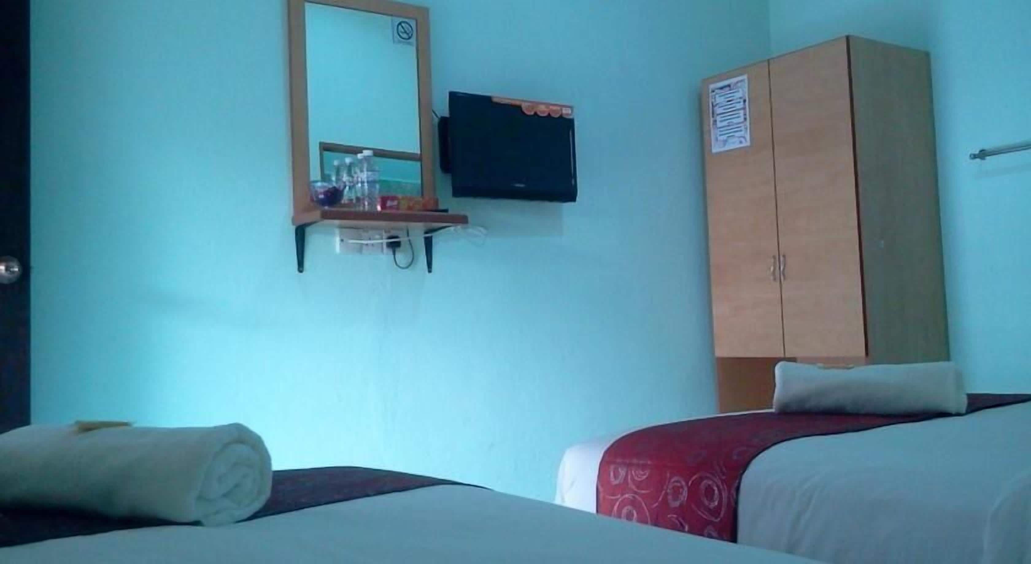 A hotel room with a bed, a wooden wardrobe, a television mounted on the wall, and a small shelf with items.