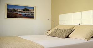 A neatly made bed with a white headboard, a beige throw blanket, and two pillows. A framed picture of a coastal scene hangs on the wall.