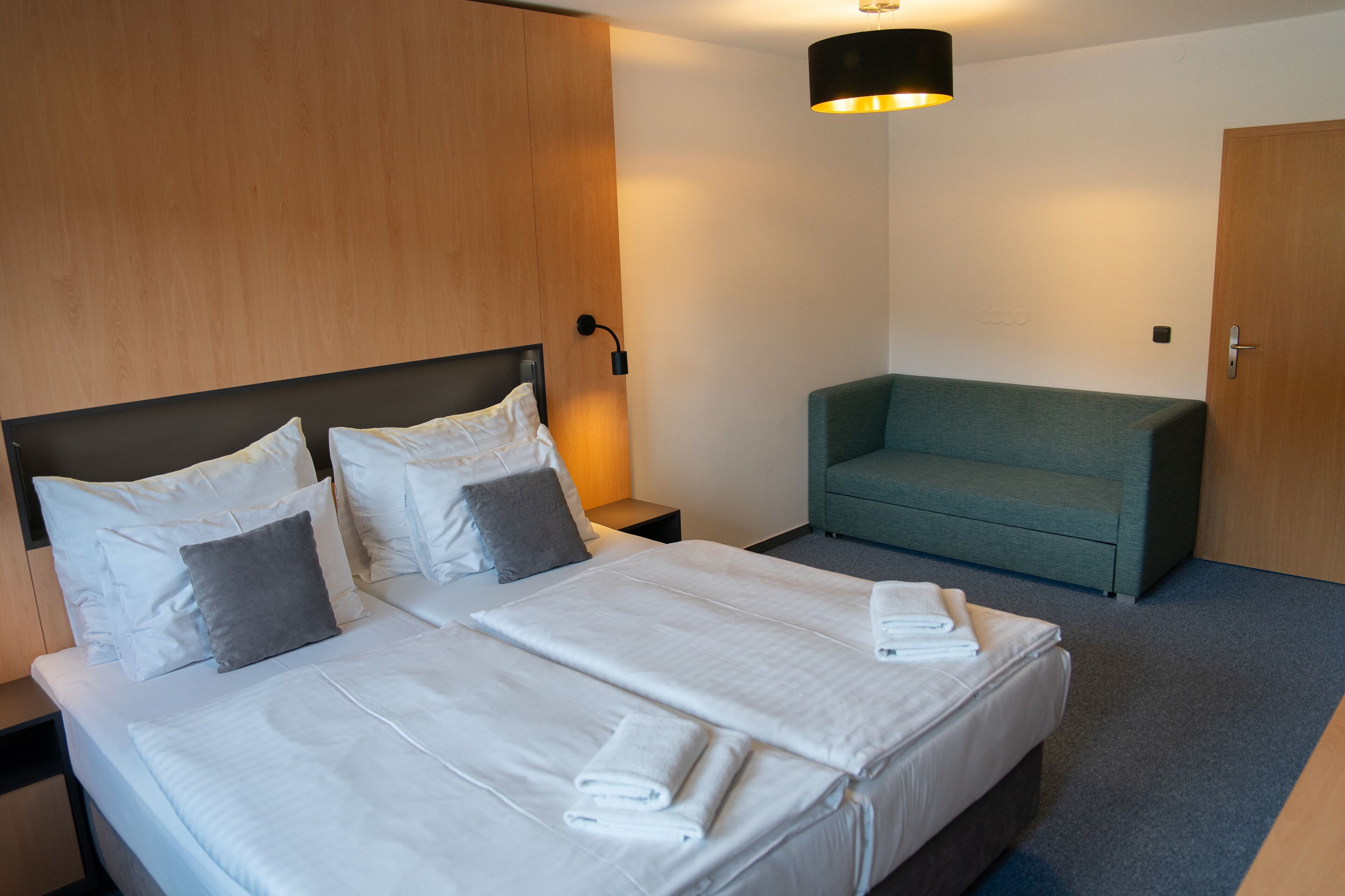 Une chambre d’hôtel avec un grand lit, deux oreillers, une table de chevet, une commode, un canapé vert et une porte.