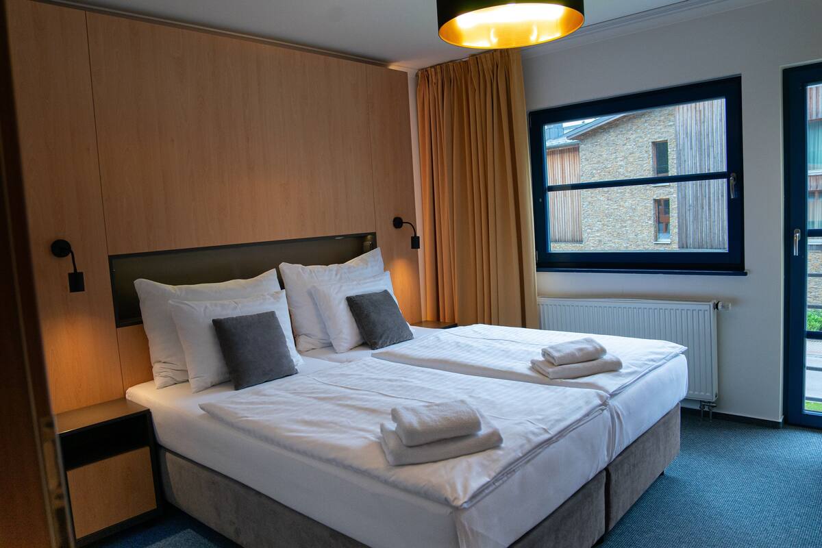 Ein Doppelbett mit weißen Laken und zwei Kissen, ein Nachttisch, ein Fenster mit Blick auf Gebäude und eine Heizung.