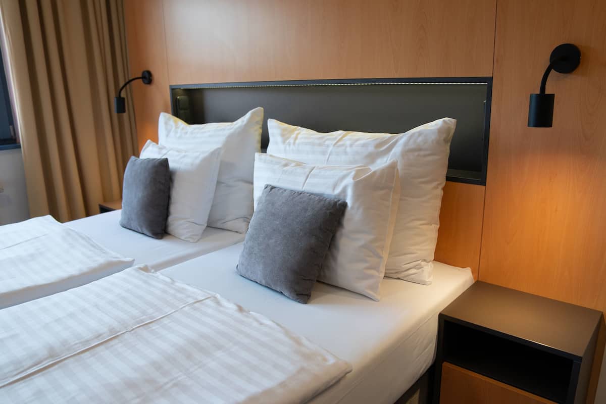 Ein Hotelzimmer mit zwei Betten, jeweils mit weißer Bettwäsche und grauen Kissen. An beiden Seiten der Betten befinden sich jeweils zwei Wandleuchten.