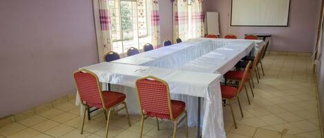 Un salón de conferencias con una larga mesa, sillas, una pantalla de proyector y ventanas con cortinas.
