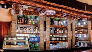 Un bar con estantes llenos de botellas de licor y vasos, una caja registradora e iluminación colgante.
