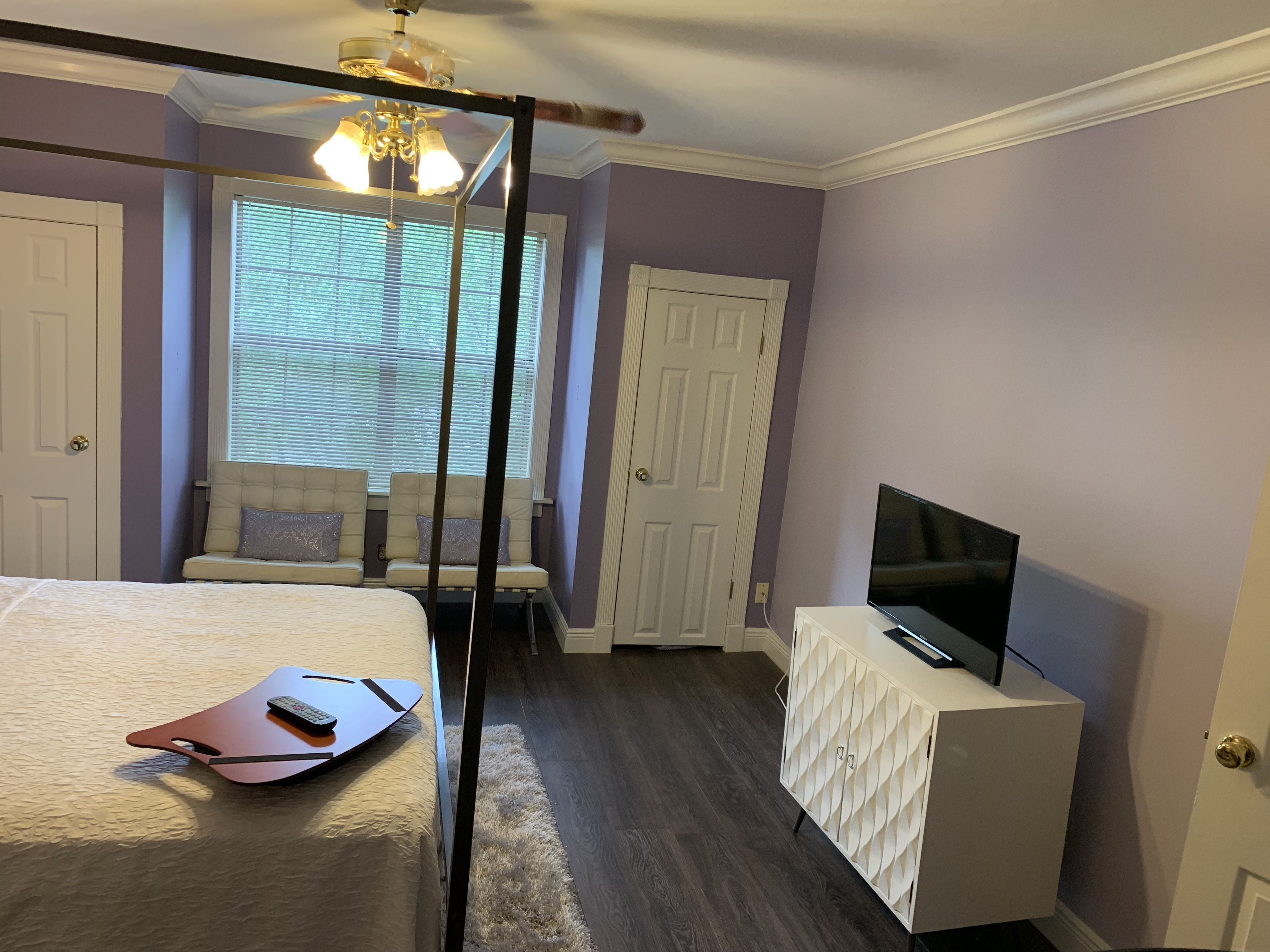 Un dormitorio con cama, televisor sobre un mueble, ventilador de techo y ventana con persianas.