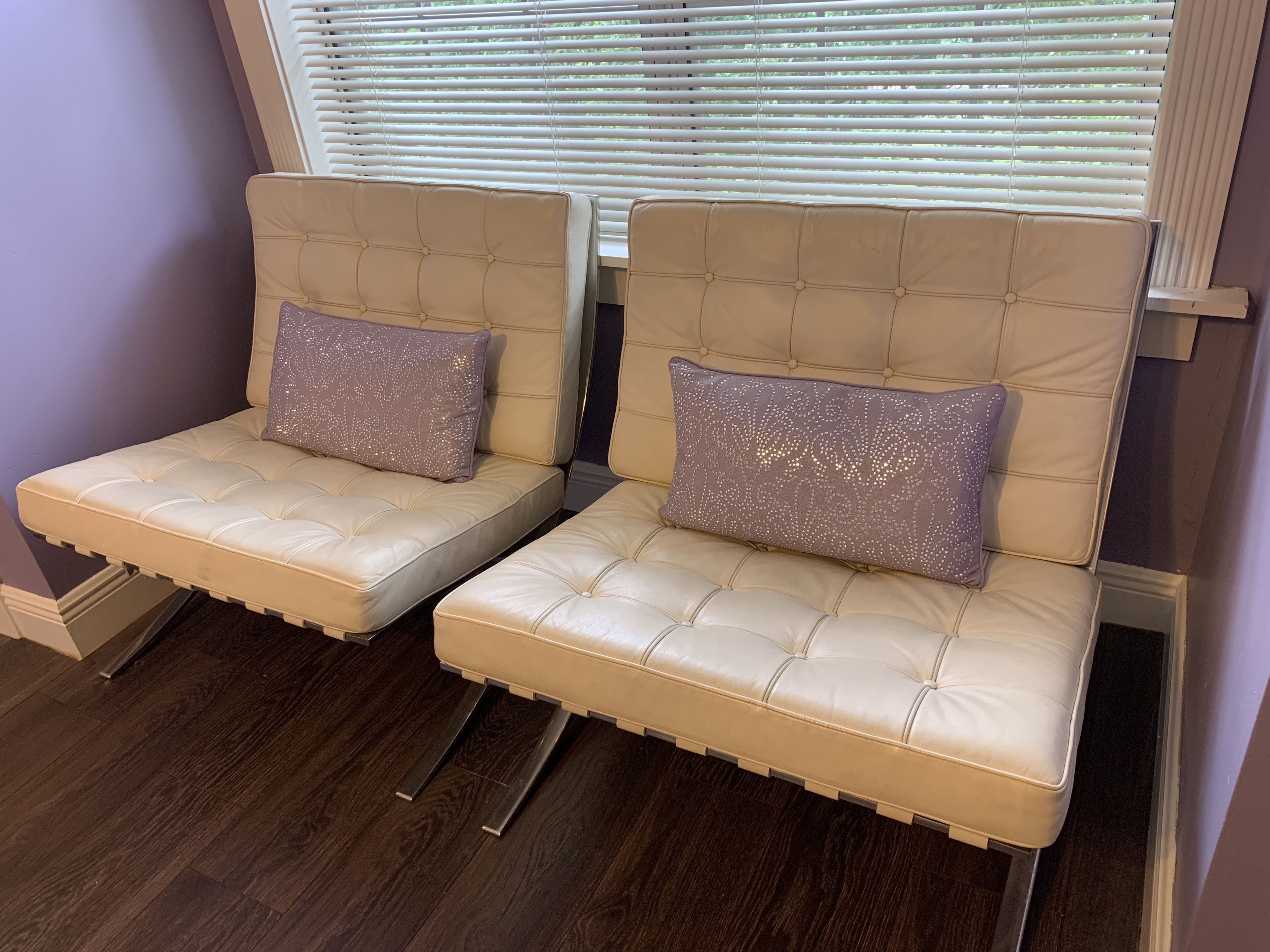 Dos sillones beige con reposabrazos capitonados y almohadones decorativos, ubicados frente a una ventana con persianas.