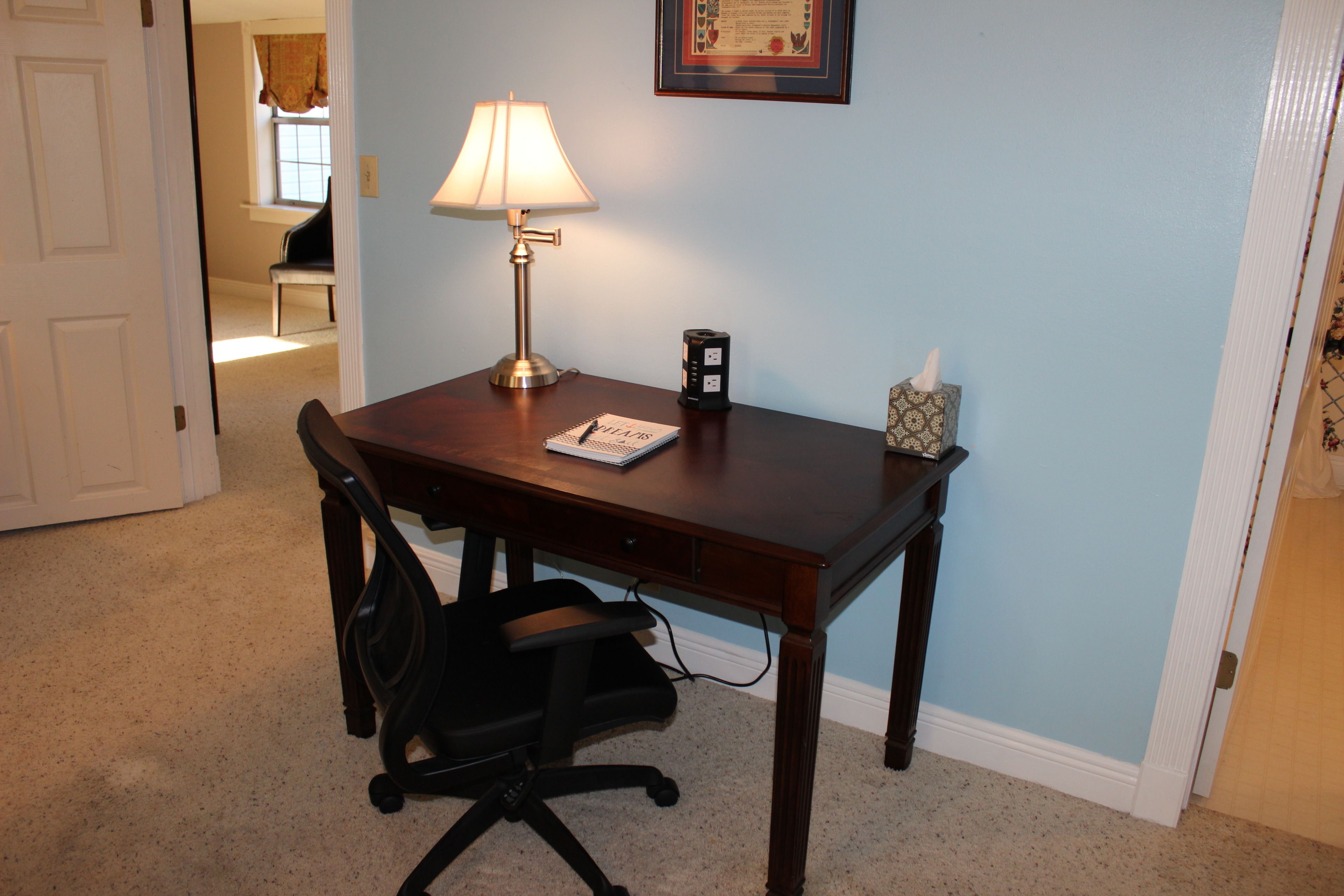 A room with a wooden desk, a black office chair, a lamp, a tissue box, and a framed certificate on the wall.