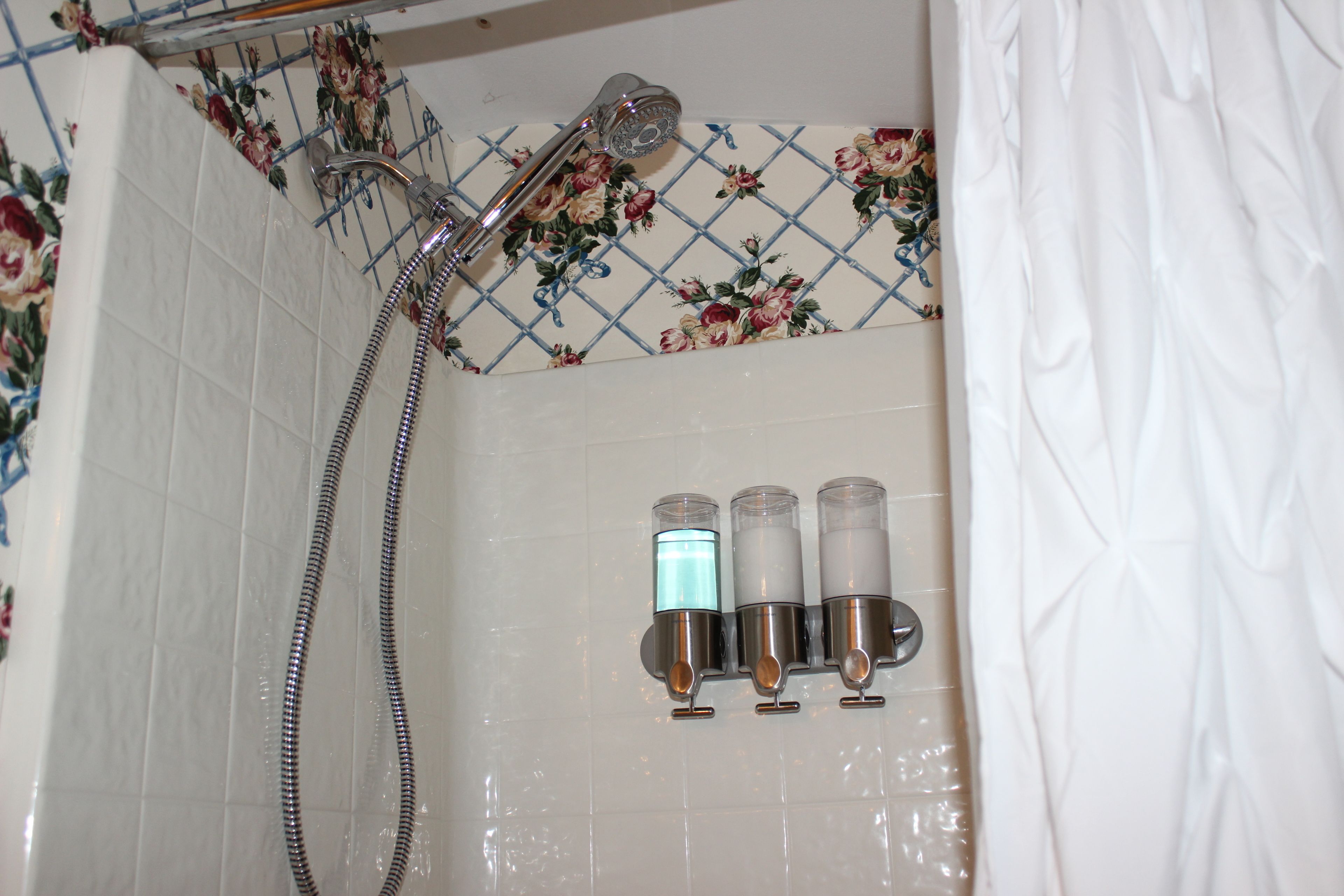 Een douche met een handdouche en een kraan op de muur. Drie flessen shampoo en conditioner staan op de muur.