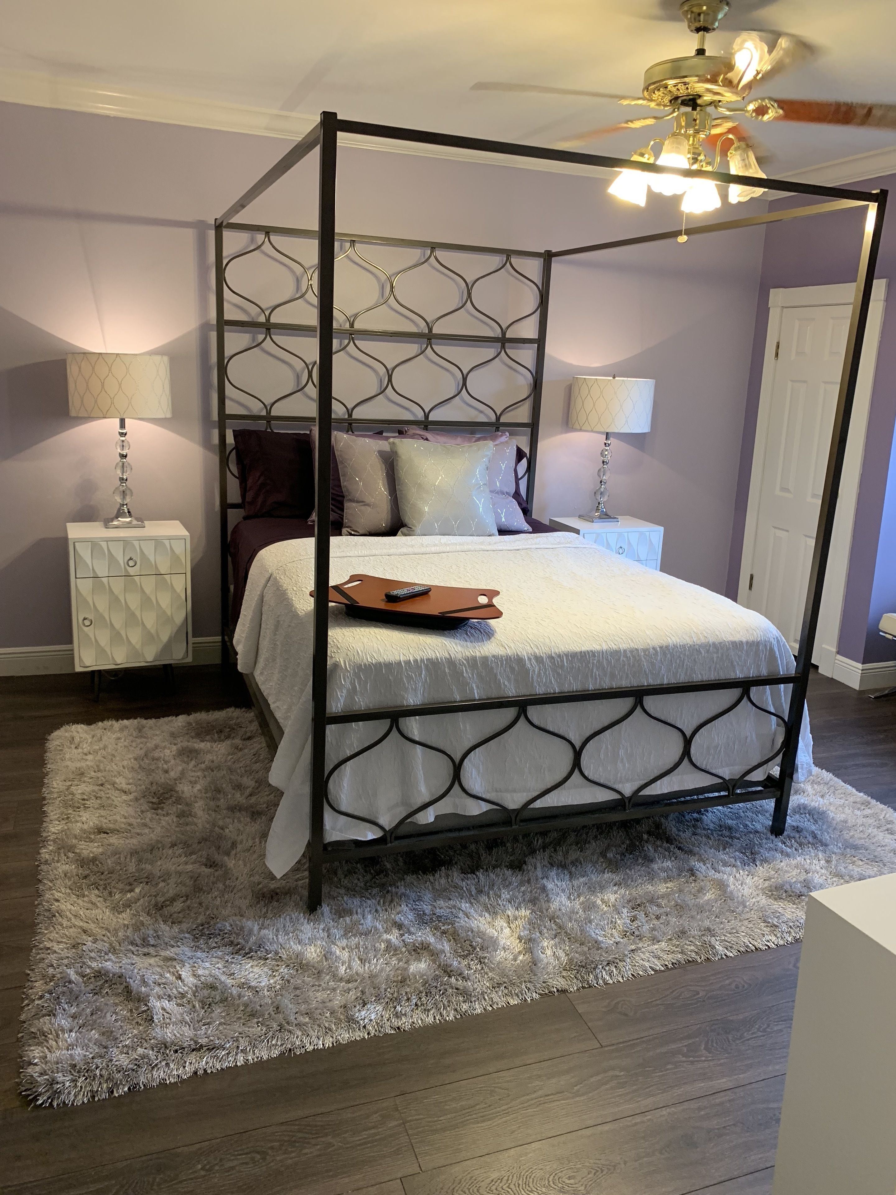 A four-poster bed with a white bedspread, two bedside tables with lamps, a fluffy rug, and a ceiling fan.
