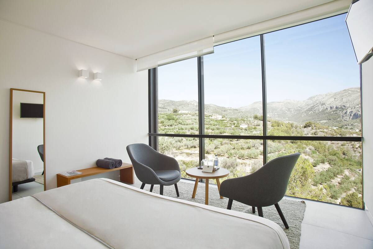 Een moderne hotelkamer met een groot bed, twee fauteuils, een bijzettafeltje en uitzicht op een woestijnlandschap door grote ramen.