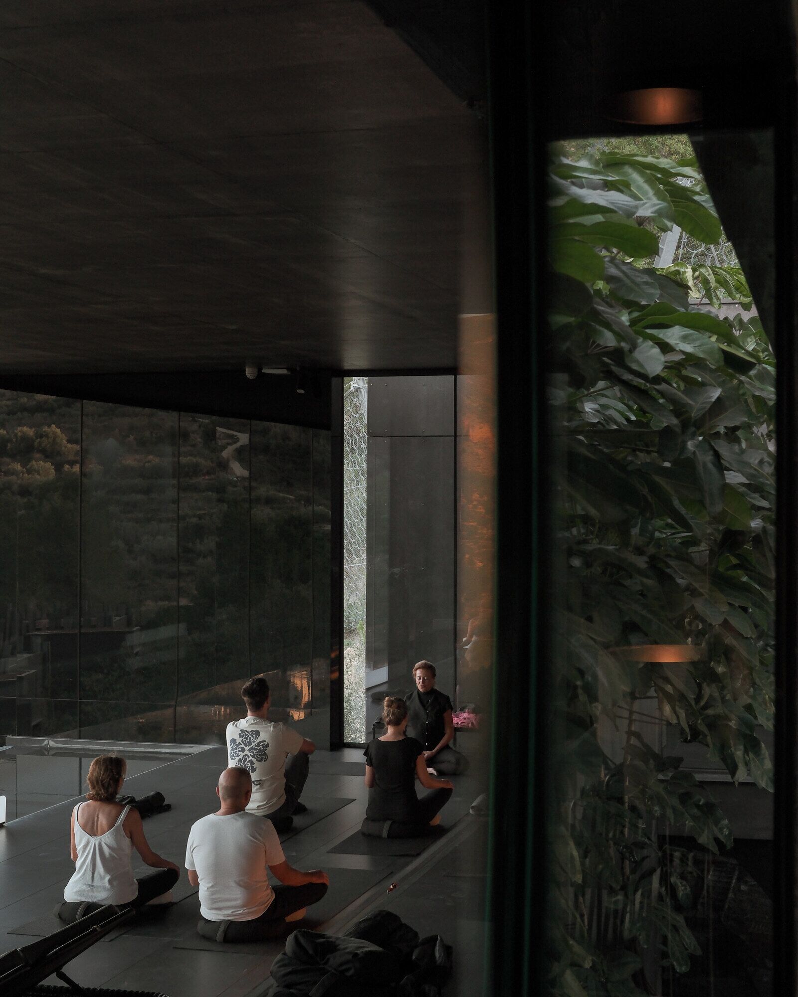A group of people sitting on mats in a modern, open space with a glass wall and a view of greenery outside.