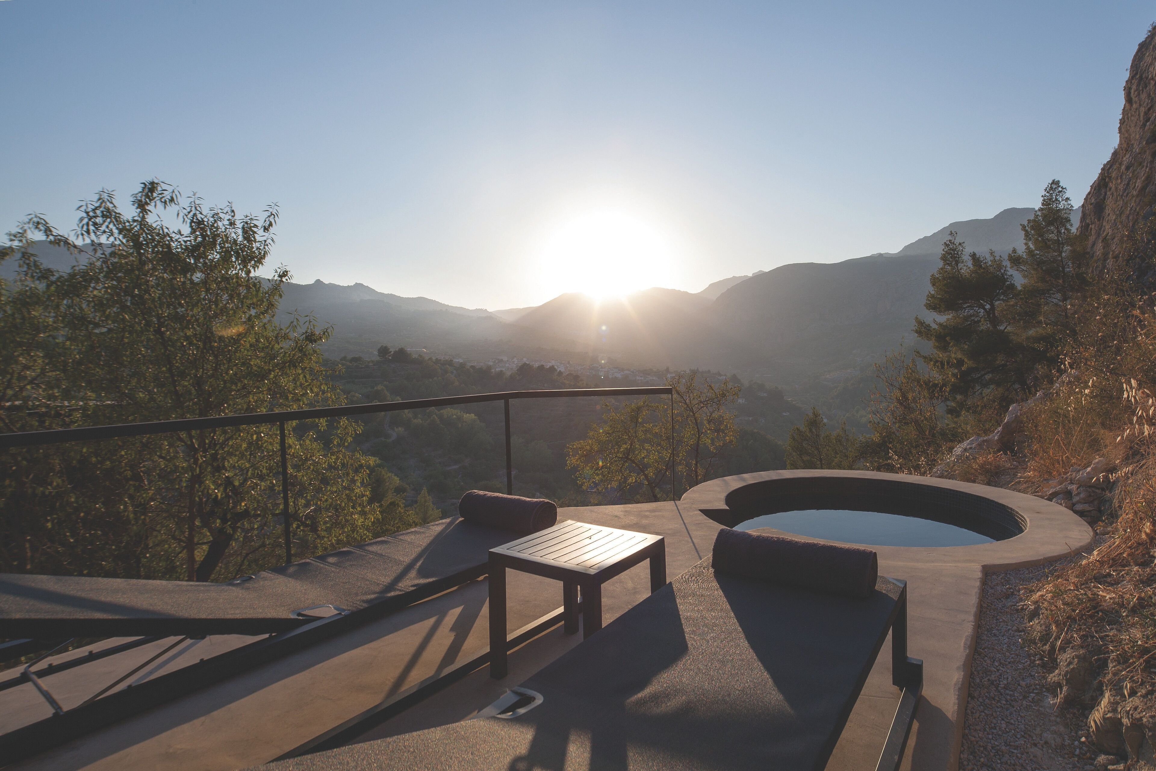 A sunlit outdoor seating area with a pool, surrounded by mountains and trees.