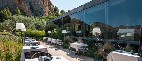 Un área de comedor al aire libre con mesas dispuestas para dos, rodeada de abundante vegetación y montañas al fondo.