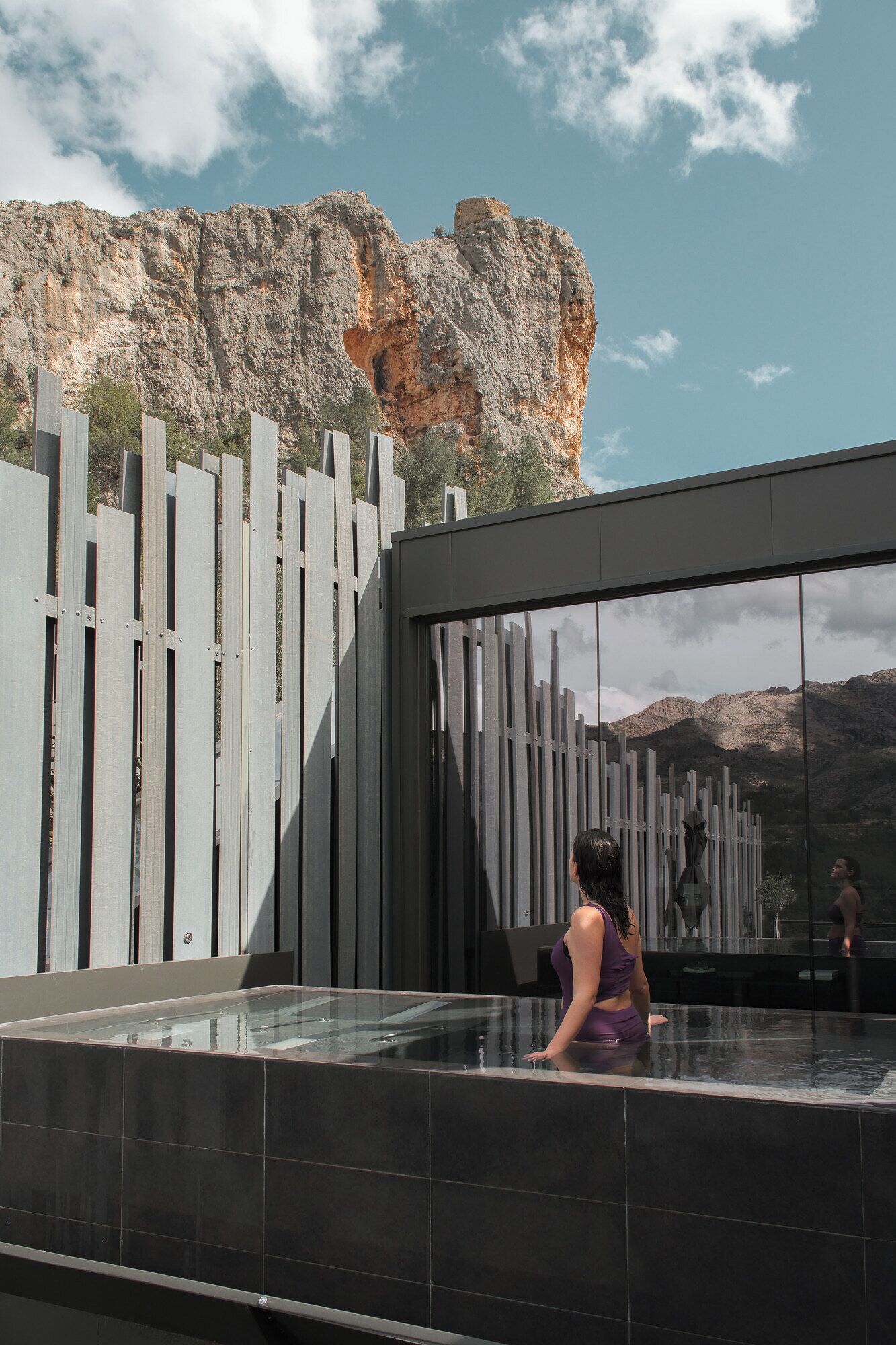 A modern outdoor pool area with a glass enclosure, a wooden fence, and a rocky mountain backdrop.