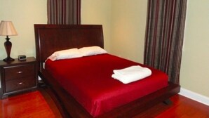 A neatly made bed with a red bedspread and white pillows, a wooden nightstand with a lamp, and striped curtains. - Choo Choo Lofts, 3 br, 2 bath, Full Kitchen - Lafayette, LA (Lafayette)
