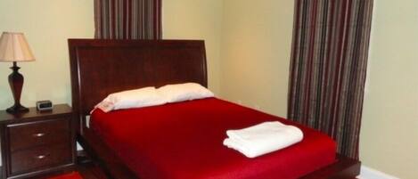 A neatly made bed with a red bedspread and white pillows, a wooden nightstand with a lamp, and striped curtains.