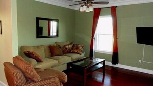 A living room with a sofa, armchair, and coffee table. There is a ceiling fan, a window with curtains, and a flat-screen TV mounted on the wall. - Choo Choo Lofts, 3 br, 2 bath, Full Kitchen - Lafayette, LA (Lafayette)