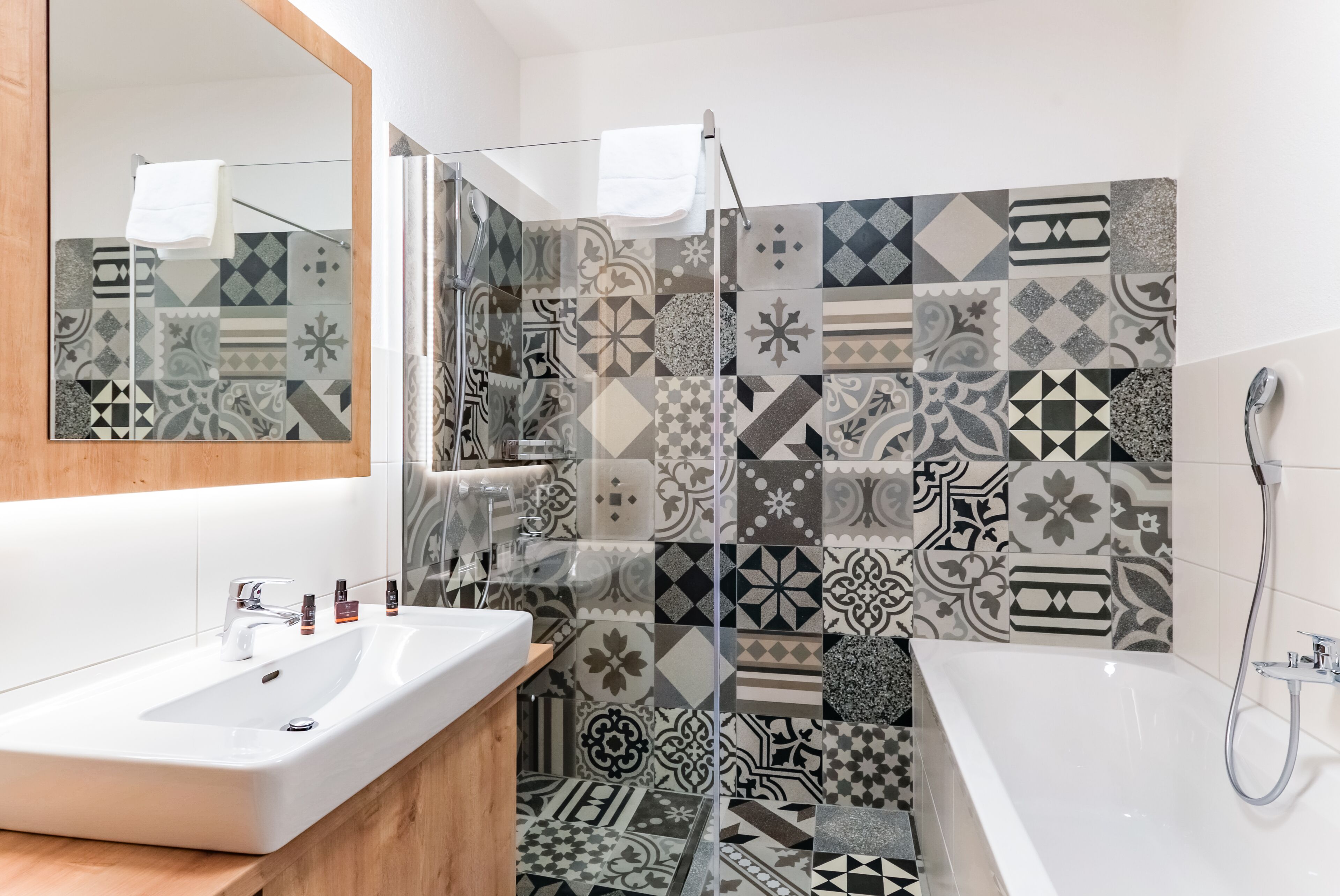 Un baño con un lavamanos blanco, un espejo, una ducha con puerta de cristal y baldosas con diseños en las paredes y el suelo.