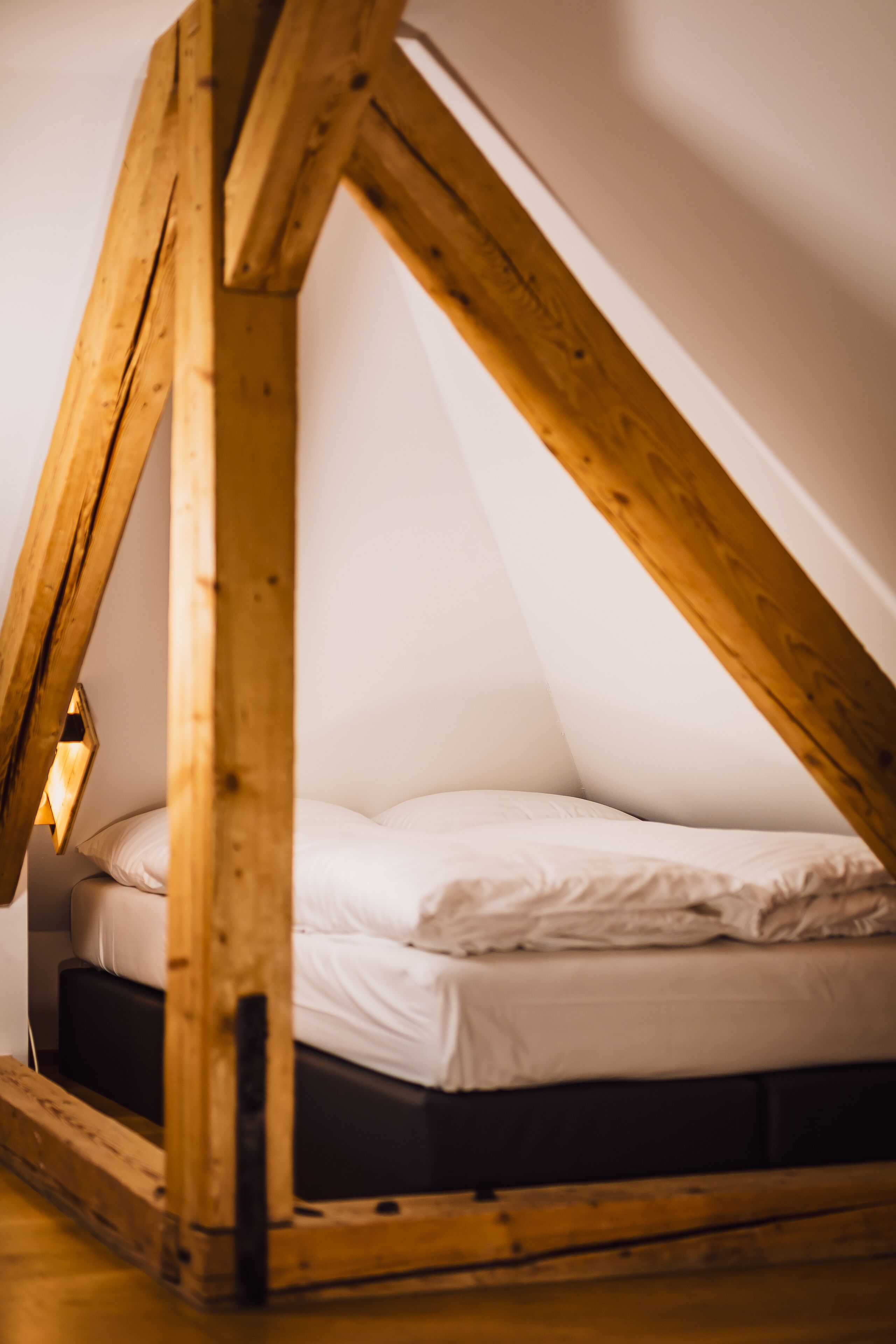 Una cama litera de madera pequeña y triangular con un colchón blanco y una mesita de noche con una lámpara.