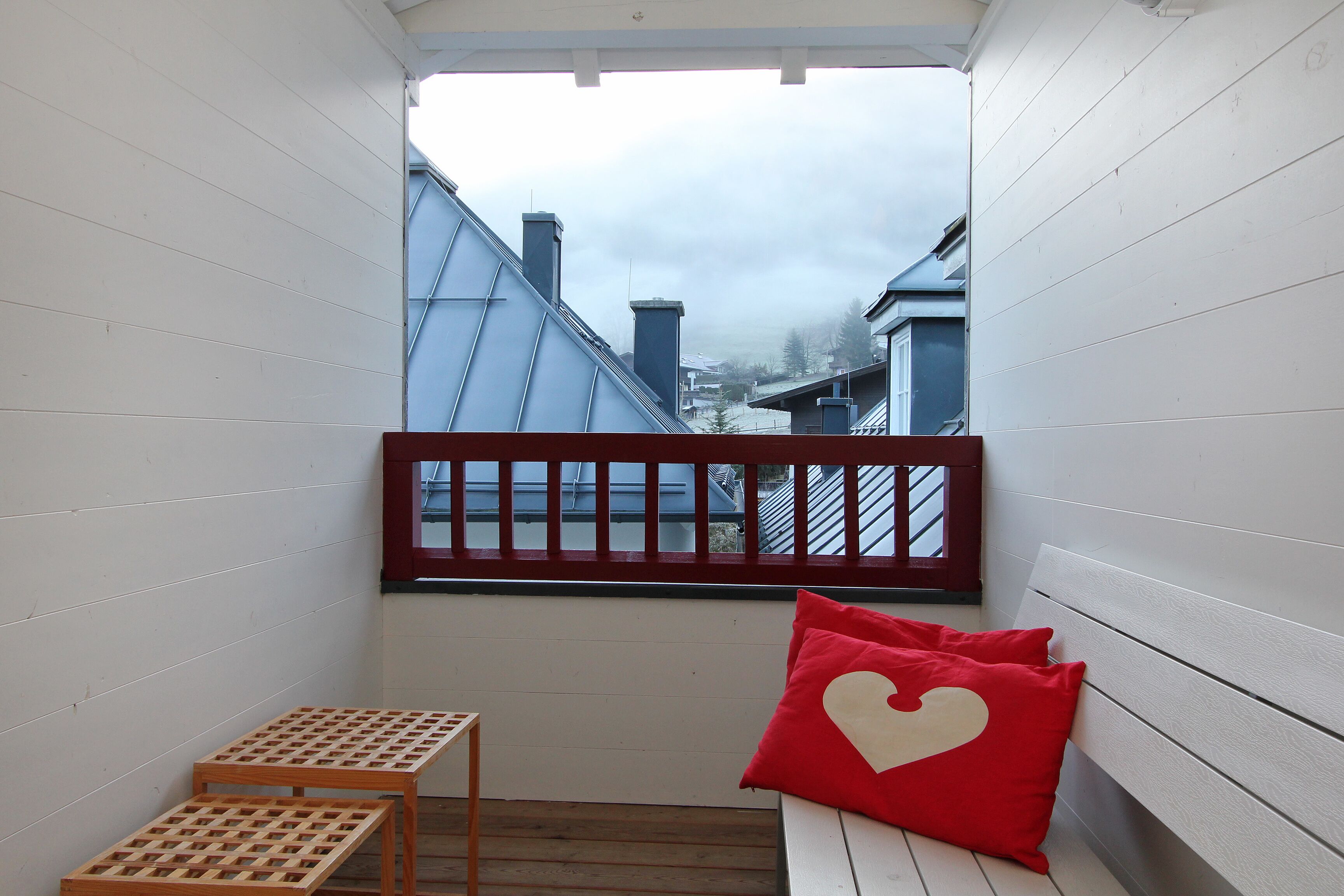 A small balcony with a wooden table, a bench, and a red heart-shaped pillow.