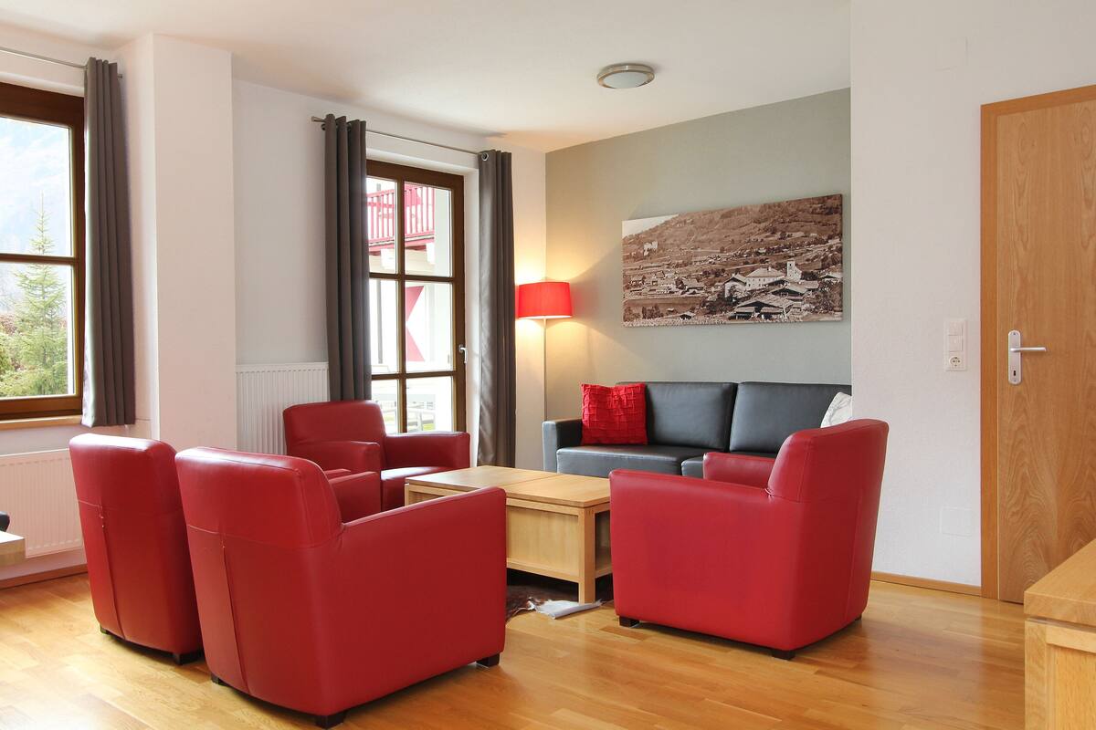 Sala de estar moderna com sofás de couro vermelho, uma mesa de centro de madeira e um abajur vermelho. Uma janela grande com cortinas e vista para um edifício é visível.