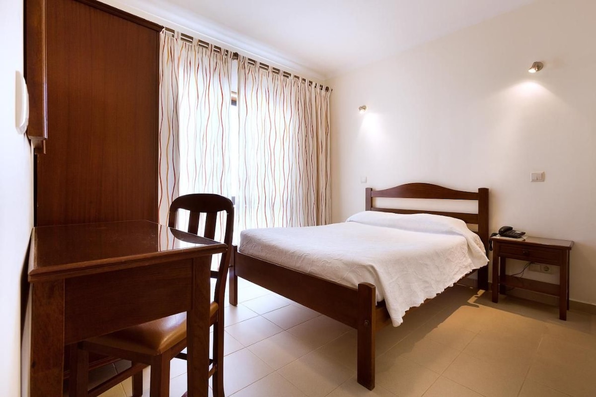 A bedroom with a wooden bed, a desk, a chair, and a bedside table.