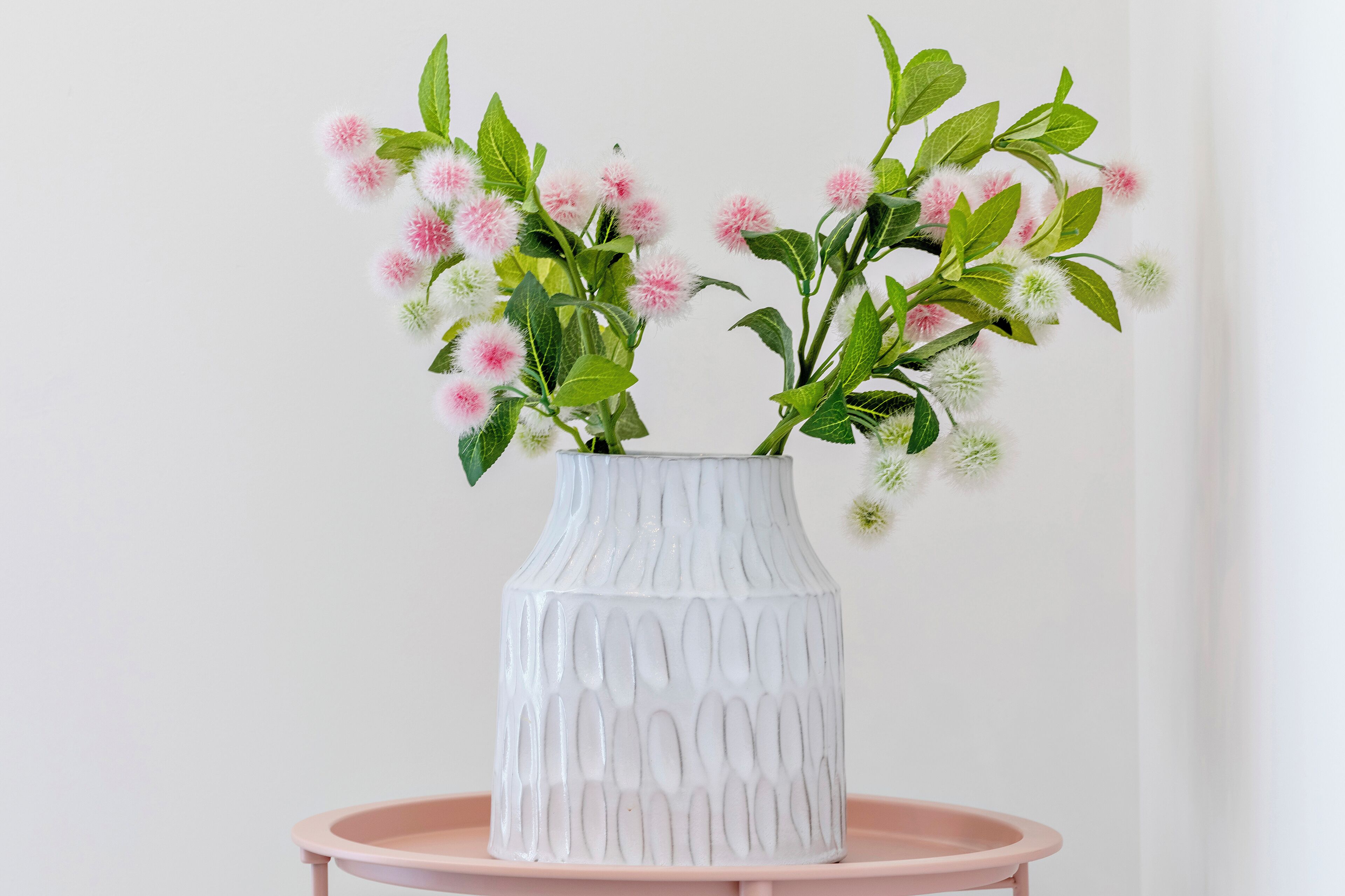 A white vase with a textured pattern containing pink and white flowers on a pink table against a white wall.