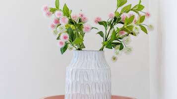 A white vase with a textured pattern containing pink and white flowers on a pink table against a white wall.