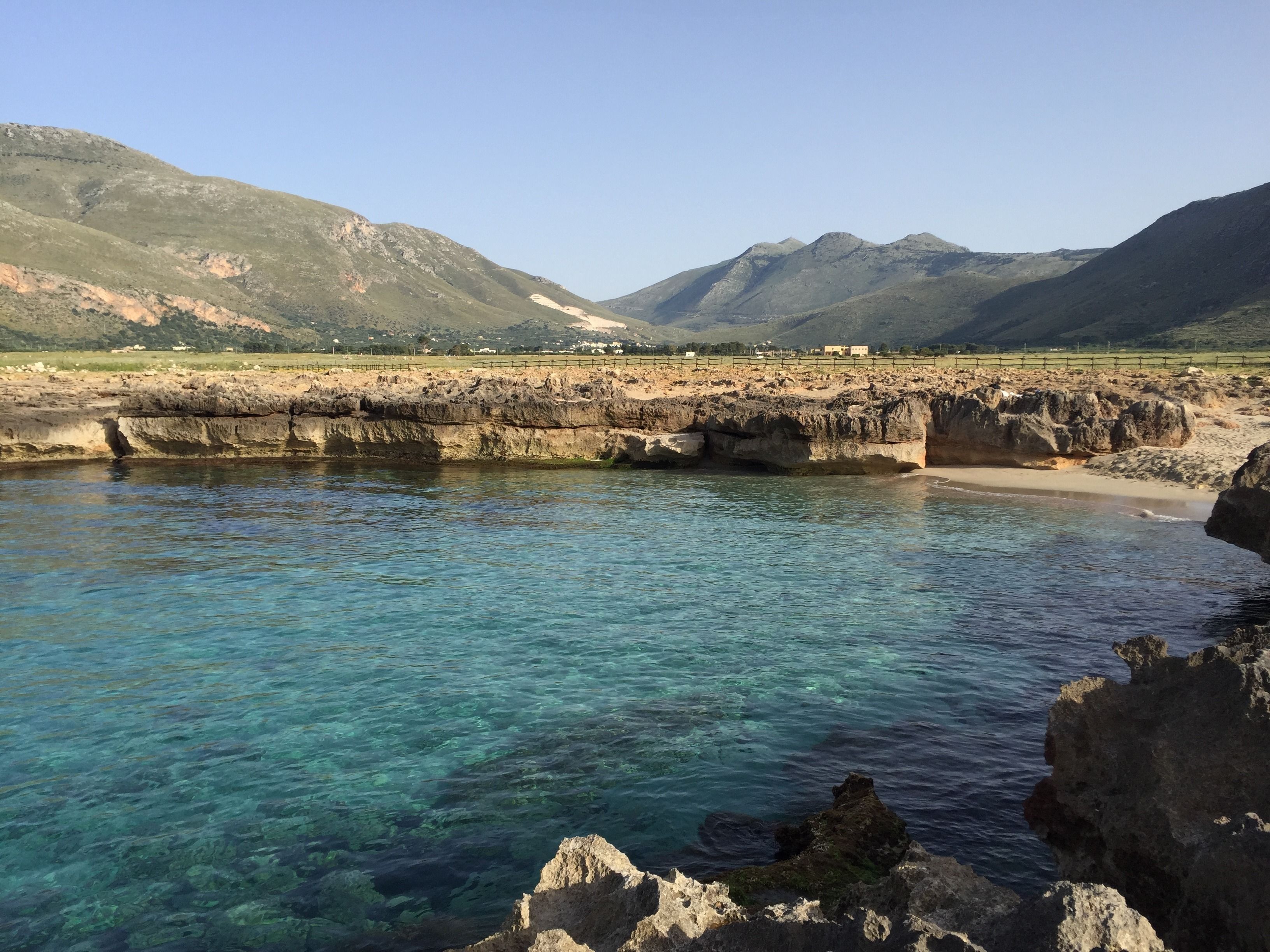 A coastal landscape with clear waters, rocky formations, and distant mountains.