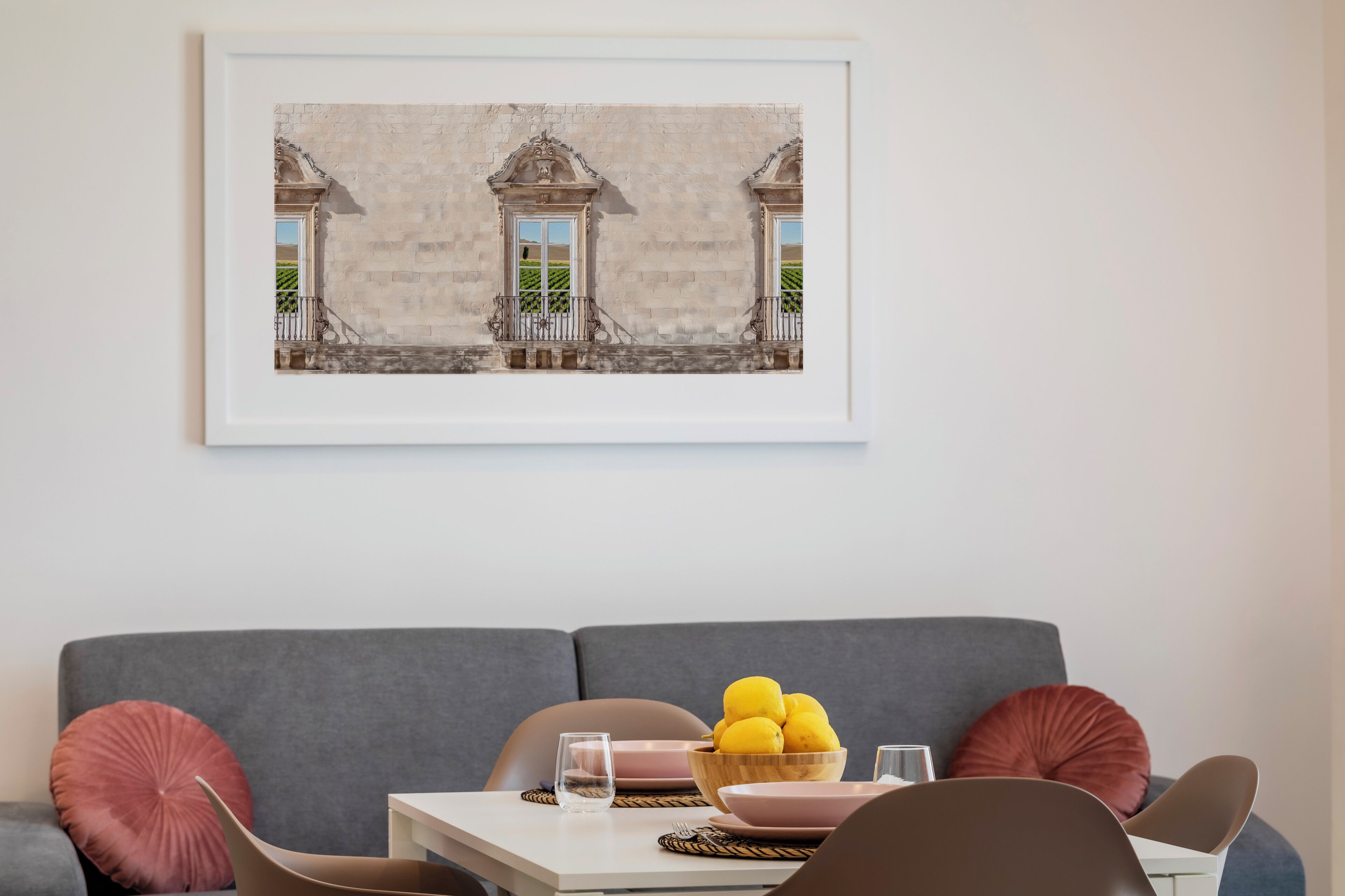 A modern living room with a grey sofa, a small dining table with a bowl of lemons, and a framed picture of a building on the wall.