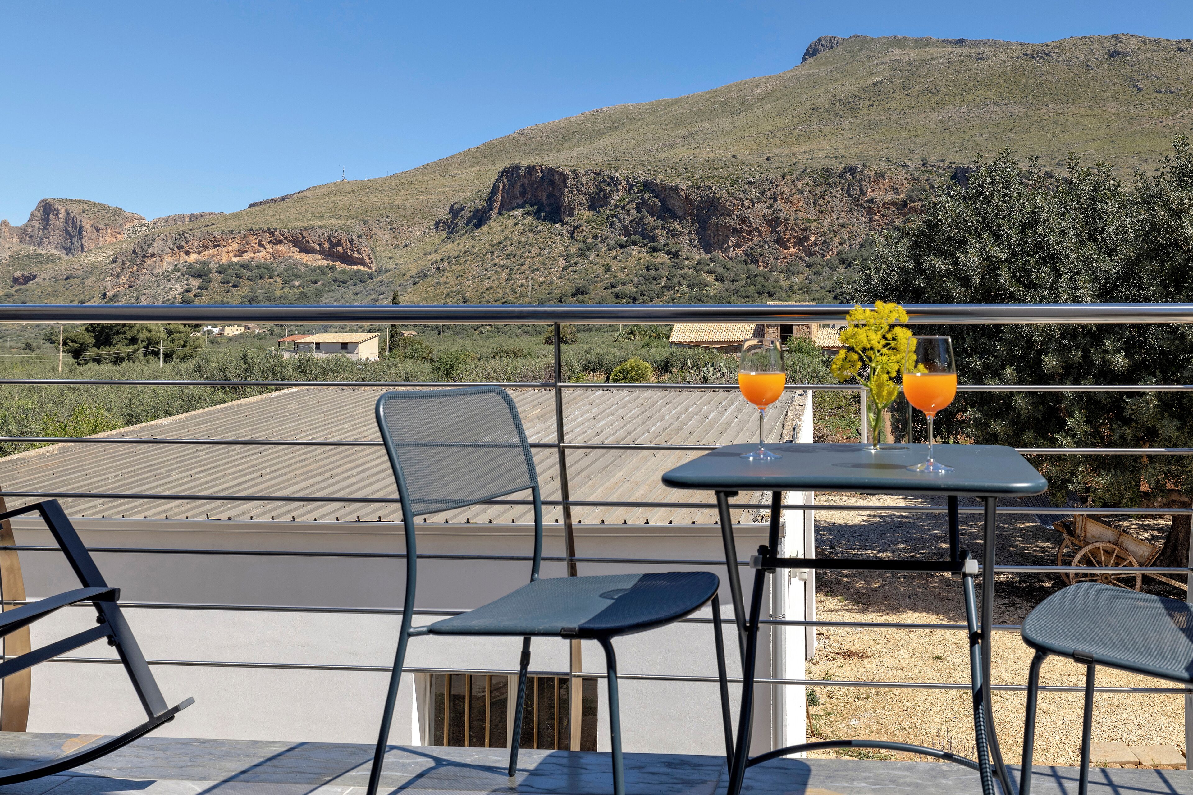 A balcony with two chairs and a table, a glass of orange juice, and a vase with yellow flowers.