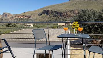 Ein Balkon mit zwei Stühlen und einem Tisch, einem Glas Orangensaft und einer Vase mit gelben Blumen.