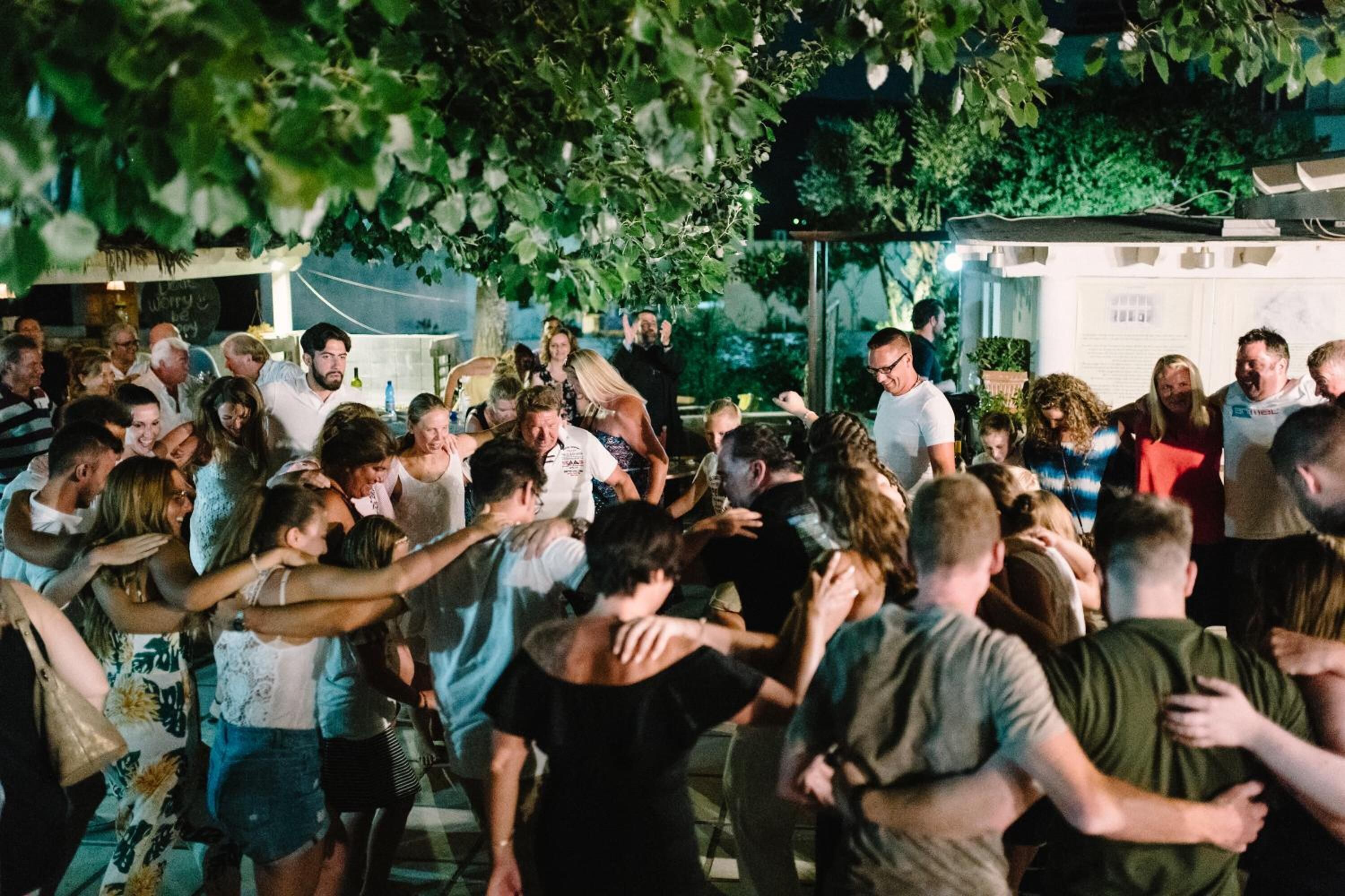 A crowded outdoor event with people dancing and socializing under a tree.