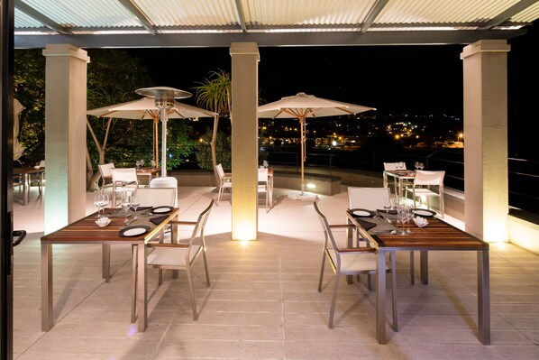 An outdoor dining area with tables and chairs, umbrellas, and a view of the city at night. - The Olive Exclusive All-Suite Hotel (Windhoek)