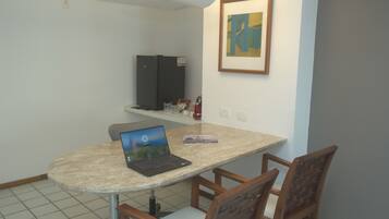 A small dining area with a round table, two chairs, a laptop, a refrigerator, a shelf with items, and a framed picture on the wall.
