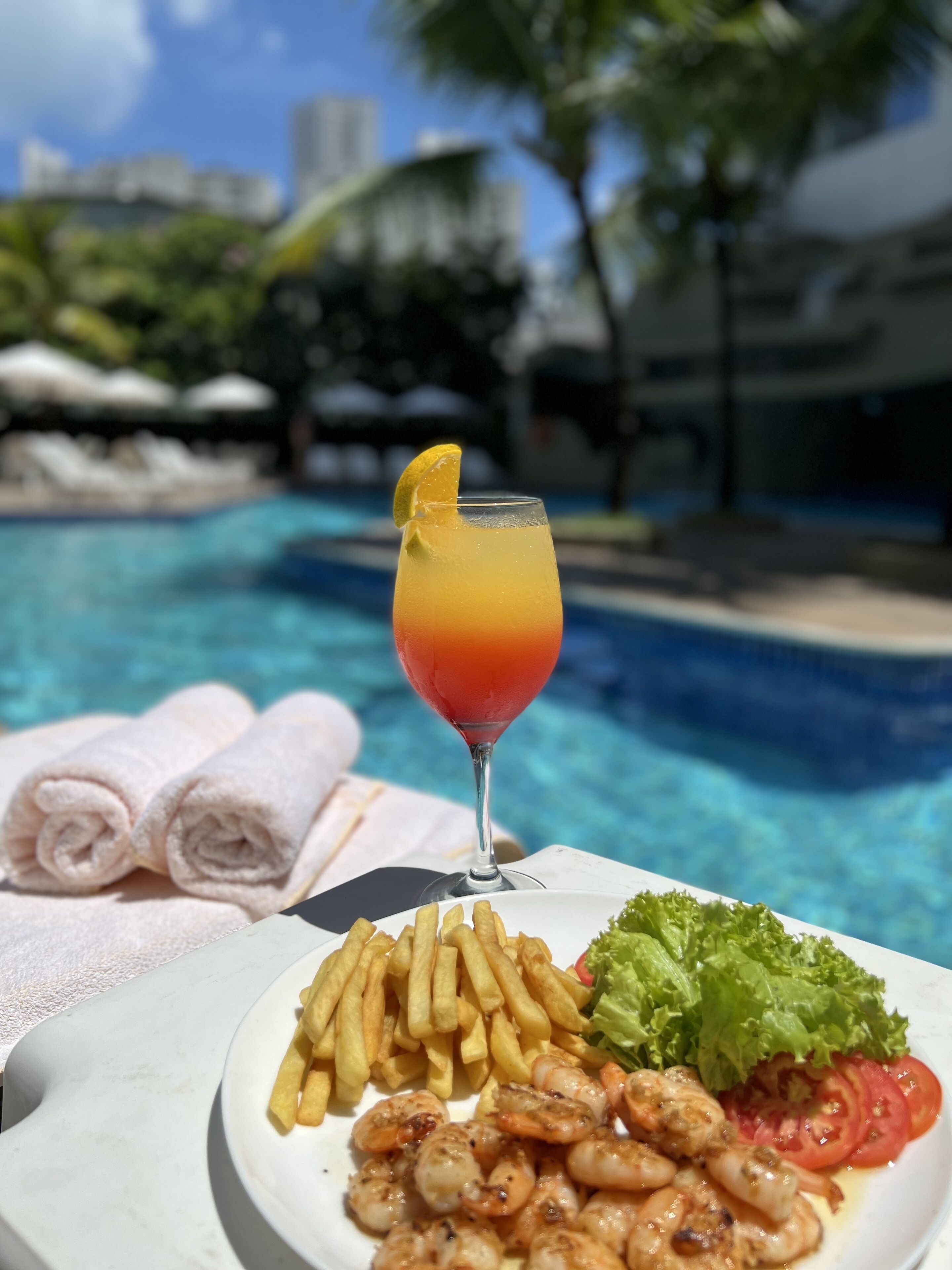 A dining table with a plate of shrimp, fries, and salad, a glass of drink, and towels by a pool.
