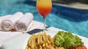 A dining table with a plate of shrimp, fries, and salad, a glass of drink, and towels by a pool.