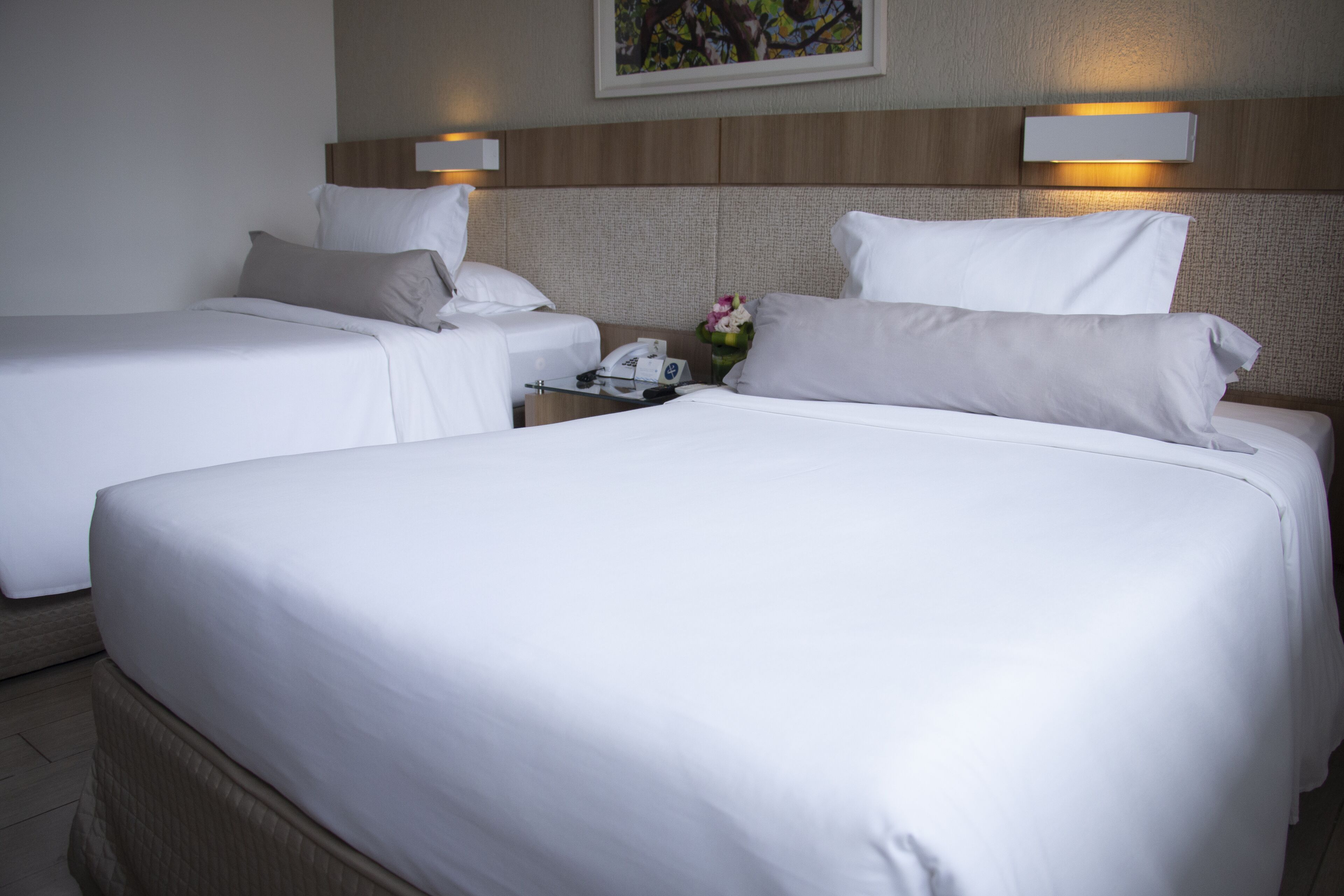 Quarto de hotel com duas camas, cada uma com roupa de cama e almofadas brancas. Há uma mesa de cabeceira com um telefone e um vaso com flores. O quarto tem uma paleta de cores neutras, com cabeceira de madeira e um quadro na parede.
