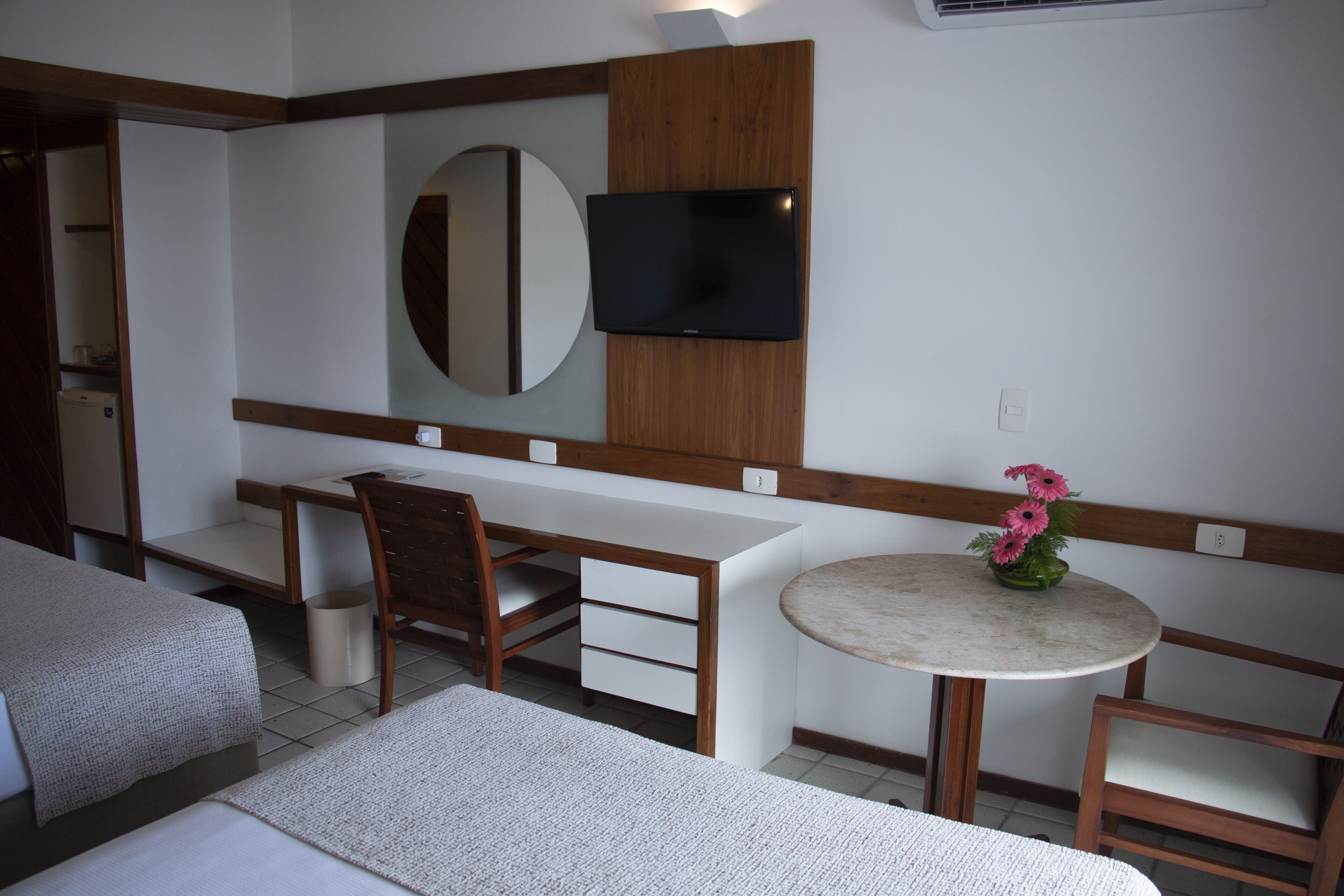 Habitación de hotel con cama, escritorio, silla, televisor montado en la pared y una pequeña mesa redonda con un florero.