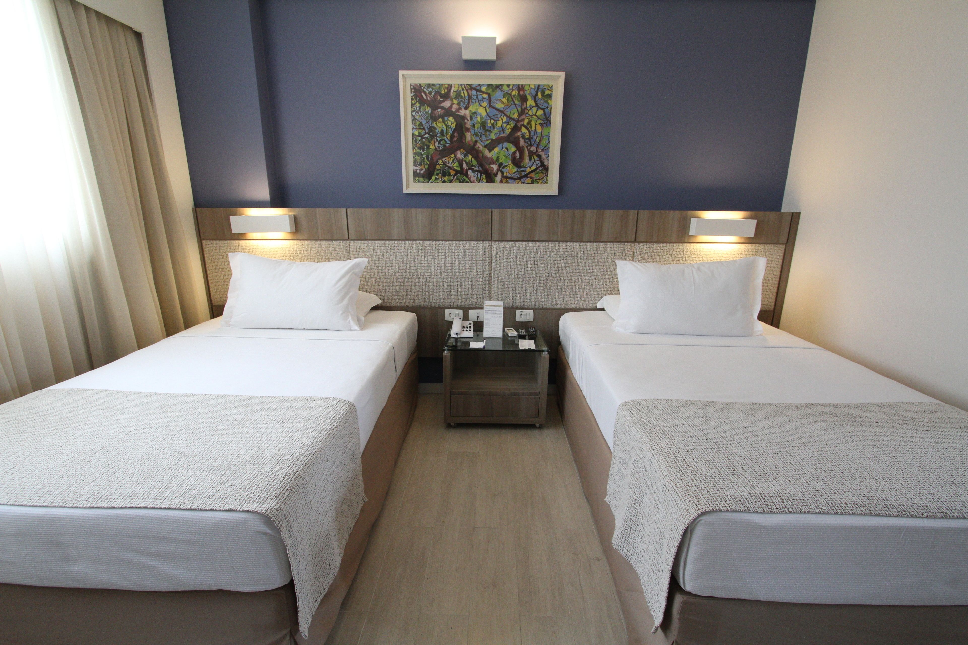 Quarto de hotel com duas camas, uma mesa de cabeceira, um quadro na parede e uma janela com cortinas.