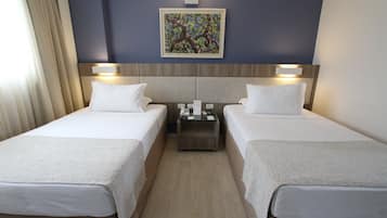 Quarto de hotel com duas camas, uma mesa de cabeceira, um quadro na parede e uma janela com cortinas.