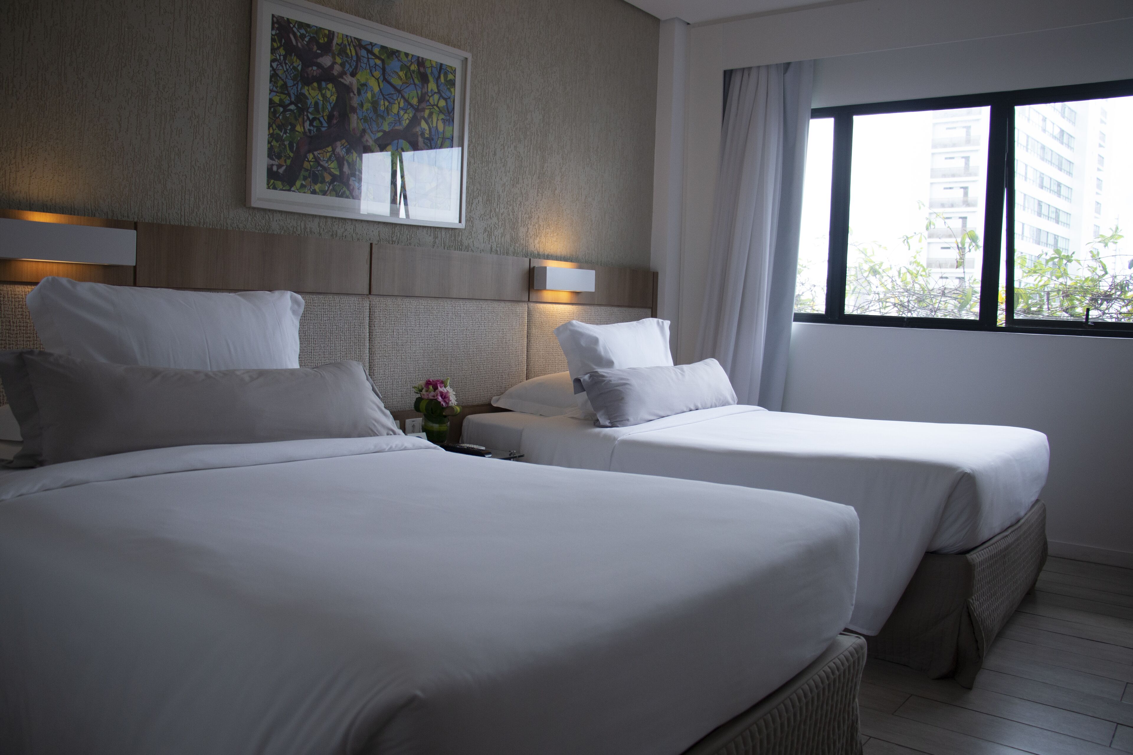A hotel room with two beds, a headboard with a light fixture, a painting on the wall, and a window with a view of buildings.