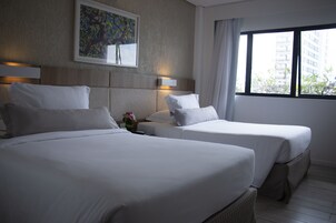 A hotel room with two beds, a headboard with a light fixture, a painting on the wall, and a window with a view of buildings.